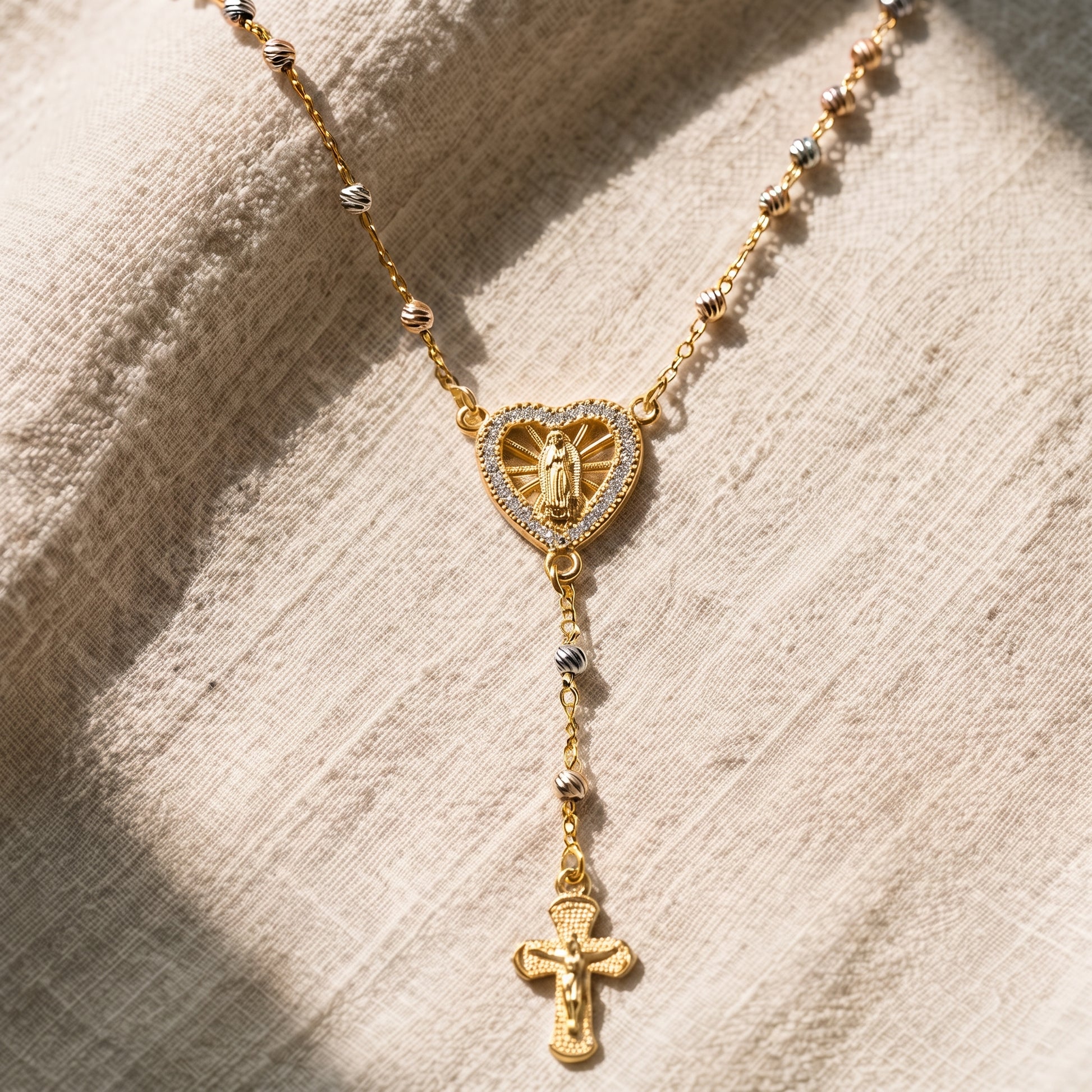 Collar rosario en tono dorado con dije de corazón de la Virgen Milagrosa engastado con circonitas, cadena de cuentas tricolor y crucifijo colgante, sobre fondo de lino natural