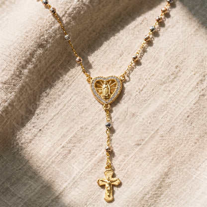 Collar rosario en tono dorado con dije de corazón de la Virgen Milagrosa engastado con circonitas, cadena de cuentas tricolor y crucifijo colgante, sobre fondo de lino natural