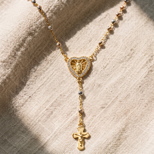 Collar rosario en tono dorado con dije de corazón de la Virgen Milagrosa engastado con circonitas, cadena de cuentas tricolor y crucifijo colgante, sobre fondo de lino natural